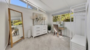 A bright, well-lit room with a large mirror, a desk, and a chair. There is a potted plant and a white dresser.