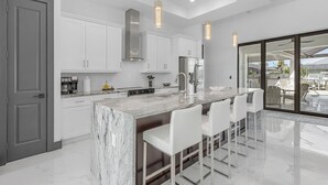 A modern kitchen with white cabinets, a marble island, and pendant lighting. - Heated Pool | Hot Tub | Game Room | Gulf Access (Cape Coral)