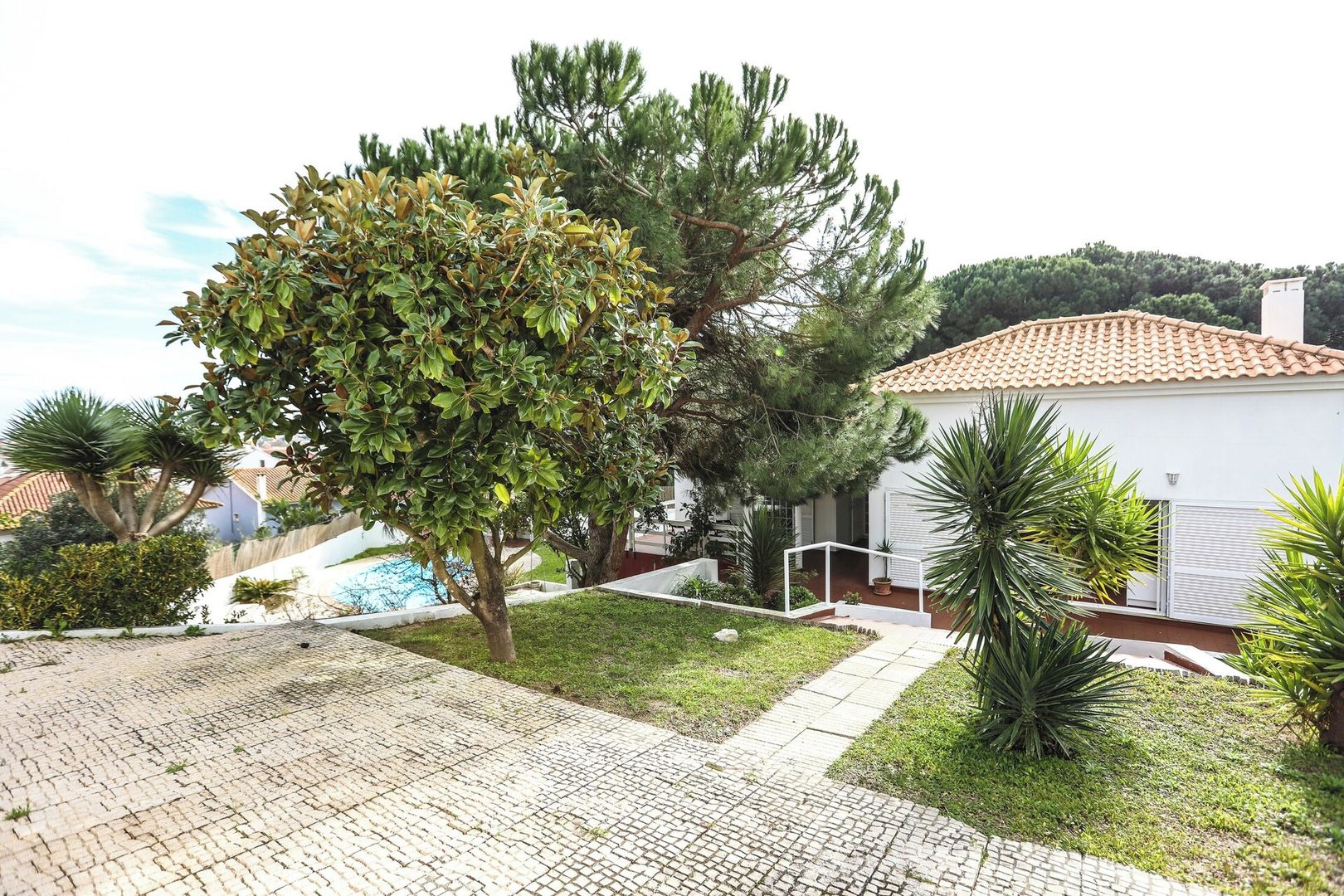 A residential property with a paved driveway, a white house with a tiled roof, and a well-maintained garden with various plants.