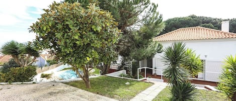 A residential property with a paved driveway, a white house with a tiled roof, and a well-maintained garden with various plants.
