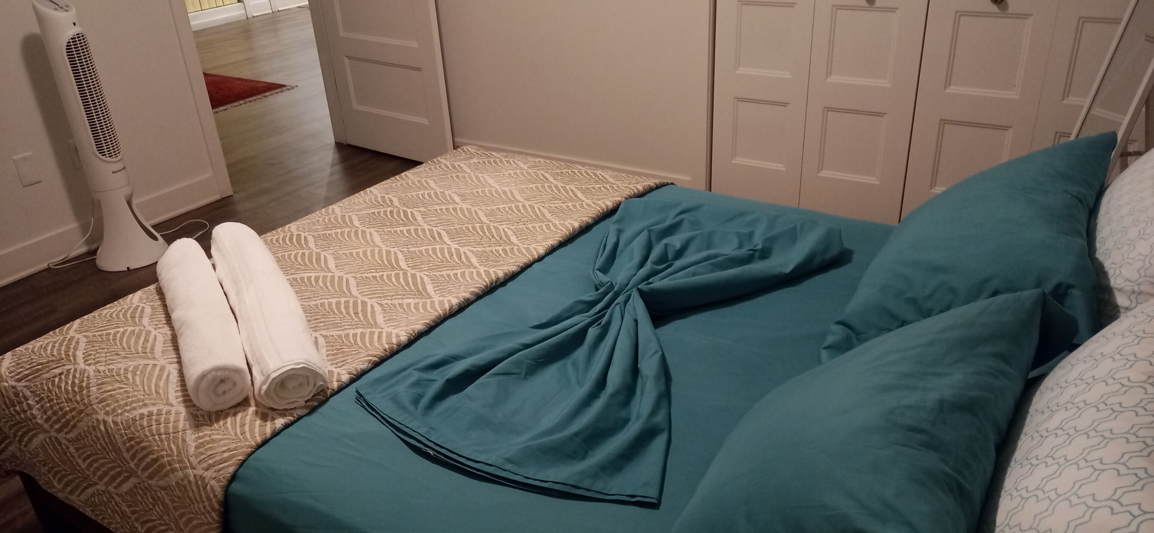 A neatly made bed with teal bedding and rolled white towels, a standing fan, and a patterned rug.
