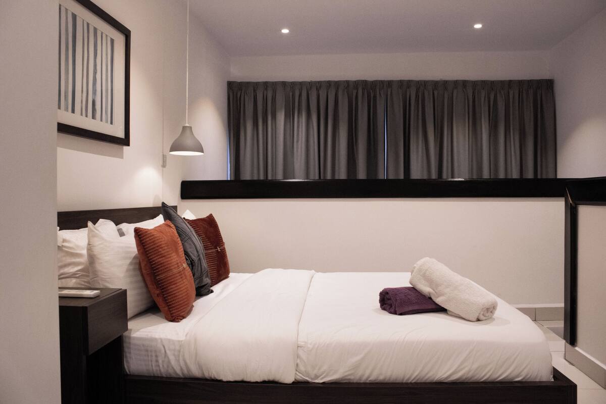 A neatly made bed with white bedding and a brown headboard, a bedside table with a lamp, and a framed picture on the wall.