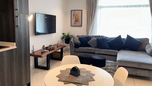 Een moderne woonkamer met een grijze bank, een ronde salontafel, een aan de muur bevestigde televisie en een groot raam met gordijnen.
