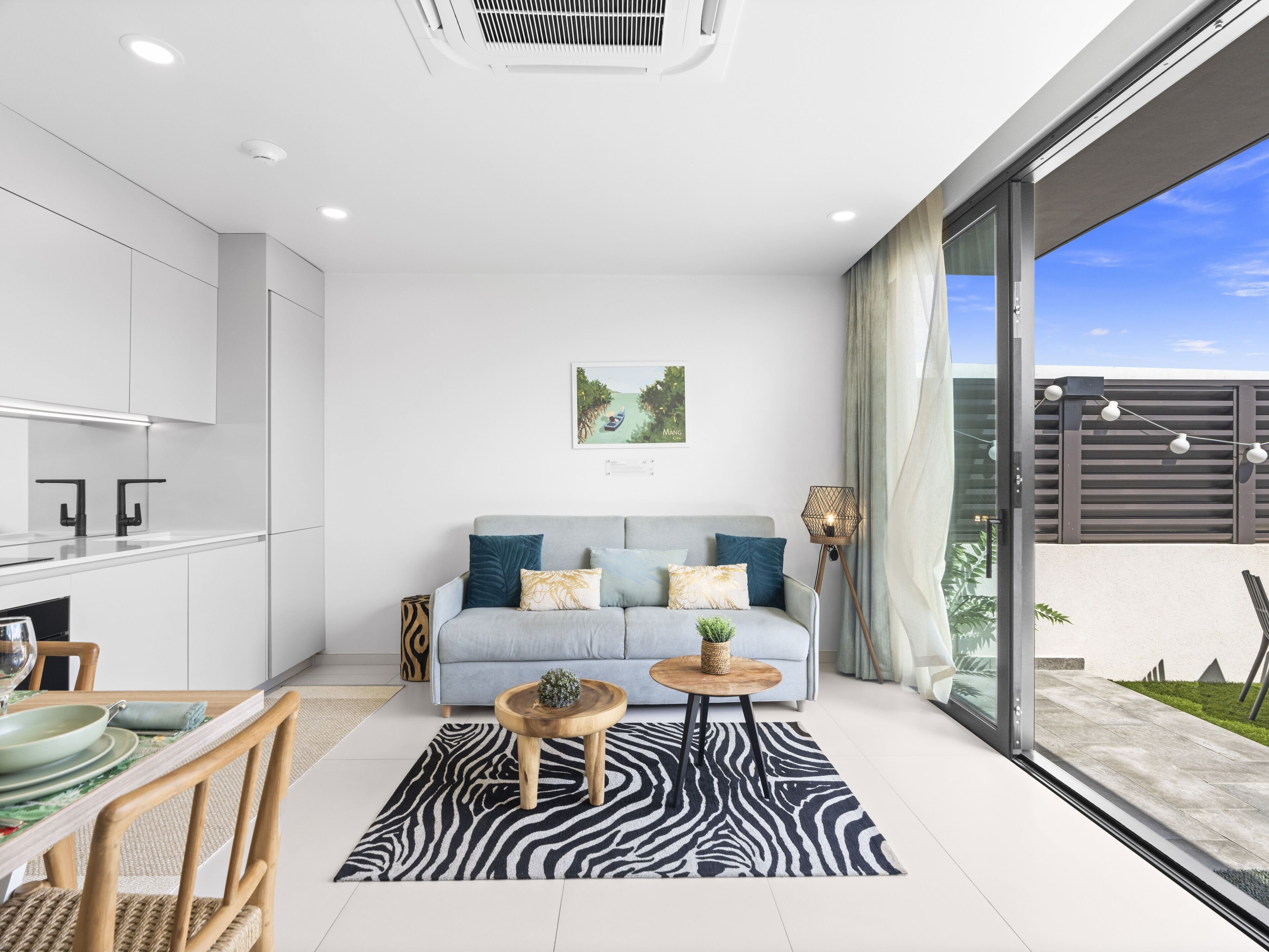 A modern living space with a kitchen, dining area, and a sofa with cushions. There is a window with curtains and a view of the outdoors.