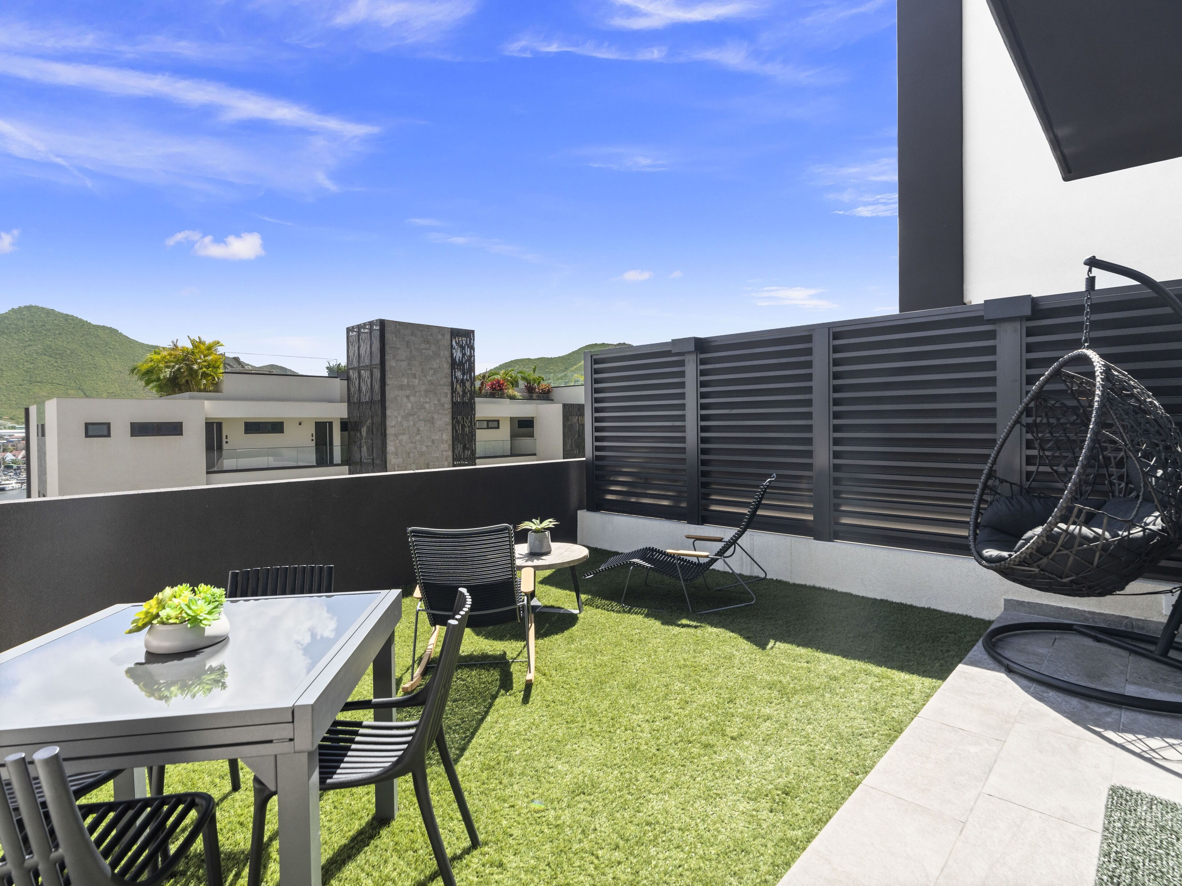 En moderne udendørs terrasse med et glasbord, metalstole, en hængestol og et lille haveområde.