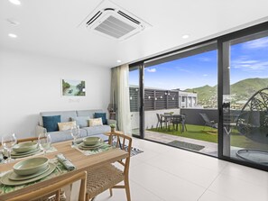 Uma sala de jantar moderna com uma mesa posta para quatro pessoas, um sofá com almofadas e uma grande porta de vidro deslizante que dá para um pátio externo com vista para a paisagem urbana e colinas.