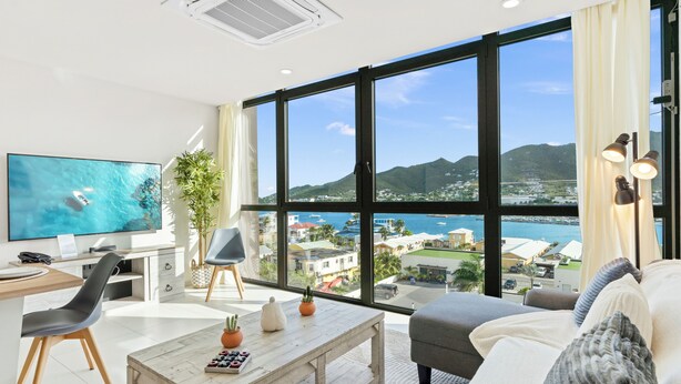 A modern living room with a large TV, a desk, and a comfortable seating area with a view of the sea and buildings. - A123 -  1 bedroom with paradise view (Sint Maarten)