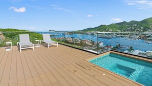 Una terraza de madera con dos sillones blancos, una piscina cubierta con vidrio y vistas a un puerto deportivo y colinas.