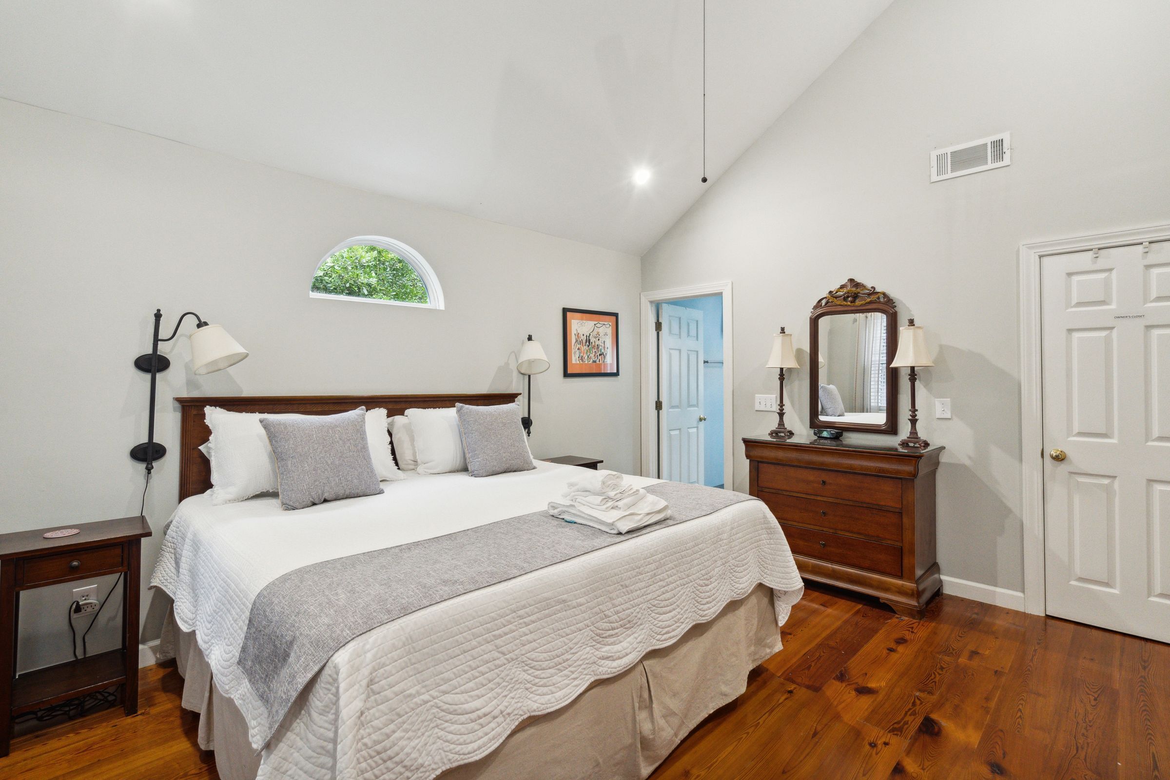 A bedroom with a large bed, a dresser, a mirror, and a window with a view of greenery.