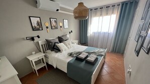 Un dormitorio ordenado con cama, mesitas de noche, una lámpara colgante y una ventana con cortinas.