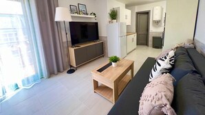 Una sala de estar moderna con un sofá, una mesa de centro, un televisor y una zona de cocina con armarios y un refrigerador.