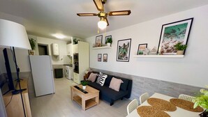 Una cocina moderna con gabinetes blancos, una zona de comedor con mesa blanca y individuales tejidos, un sofá oscuro con cojines decorativos y un ventilador de techo.
