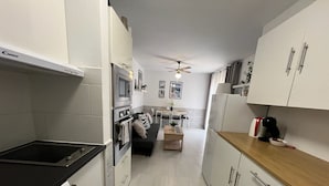Una cocina moderna con encimera negra, armarios blancos y horno empotrado. Hay una zona de comedor con sofá negro y mesa de madera.