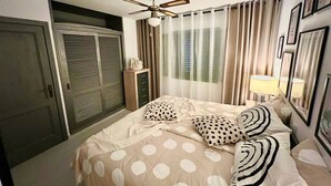 Un dormitorio con cama, ventilador de techo, ventana con cortinas, mesita de noche con lámpara y armario.