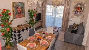 A dining area with a wooden table set for four, surrounded by chairs. A chandelier hangs above, and there are decorative plants with red flowers.