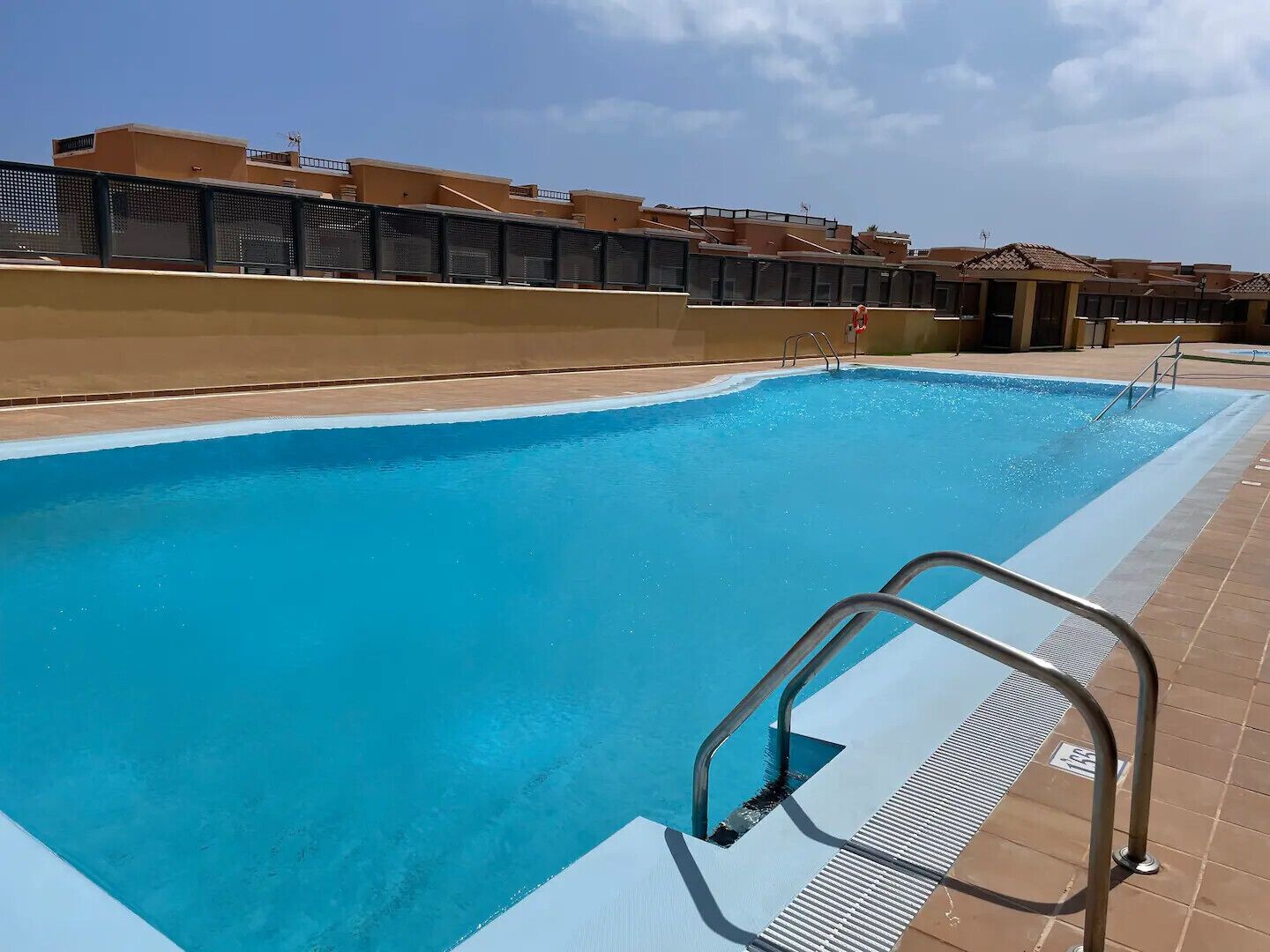 A swimming pool with a metal ladder, surrounded by a tiled deck and a low wall.
