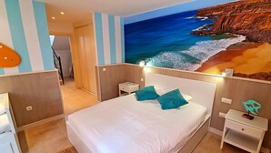 Ein Hotelzimmer mit einem Bett, zwei Nachttischen, einem Wandbild mit Strandmotiv und einer Tür zu einem anderen Raum.