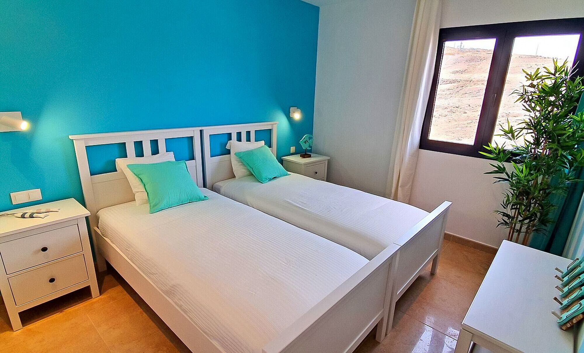 A double bed with white bedding and turquoise pillows in a room with blue walls and a window with a view of a desert landscape.