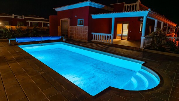Ein Swimmingpool mit blau beleuchtetem Rand, einem überdachten Sitzbereich und einem Gebäude mit roter Fassade und weißem Zierrat.