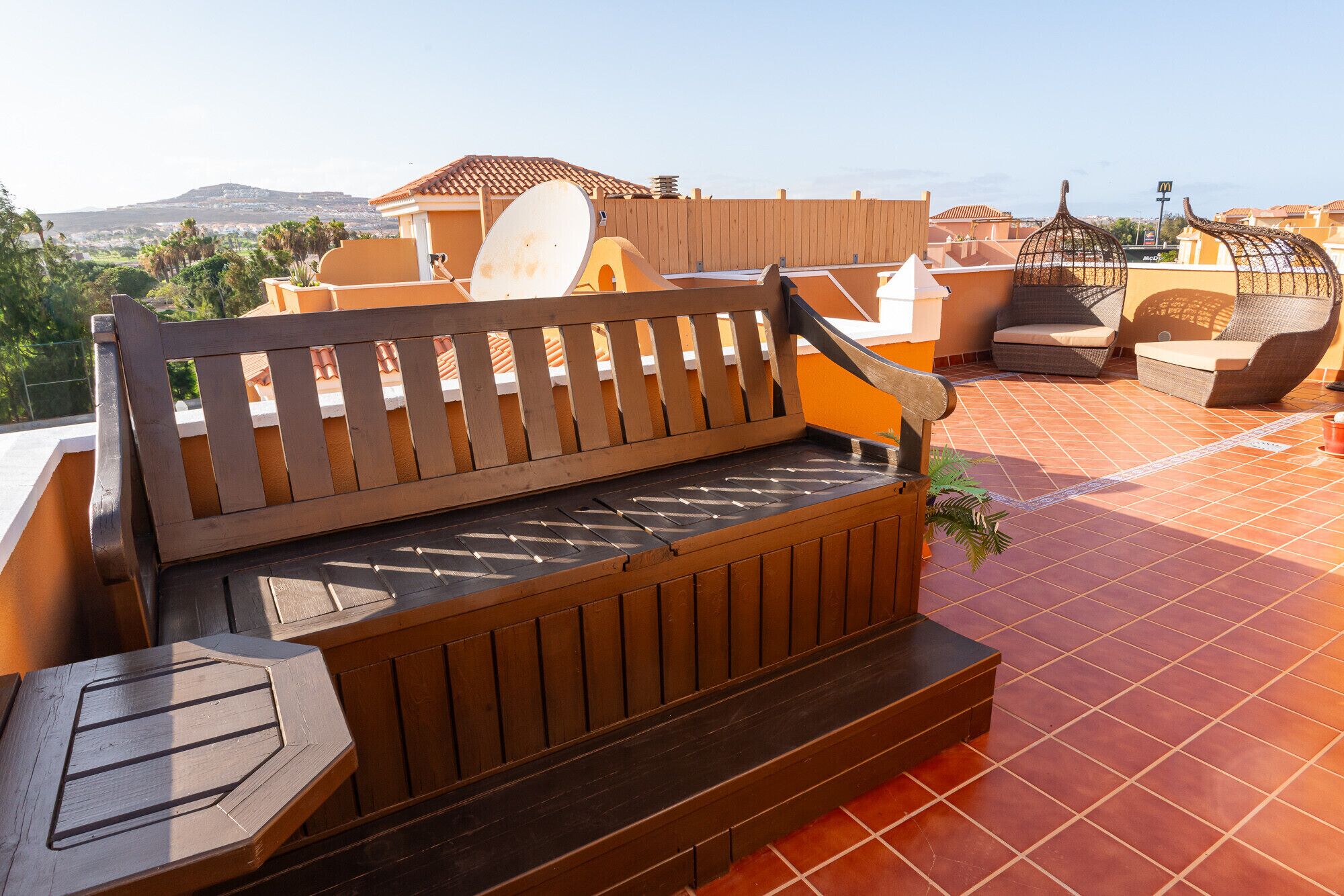 A wooden bench with a metal tray on a tiled patio with a view of buildings and a mountain in the background.