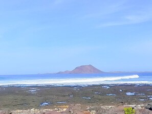 Una playa con primer plano rocoso, olas acercándose a la orilla y una montaña en la distancia.