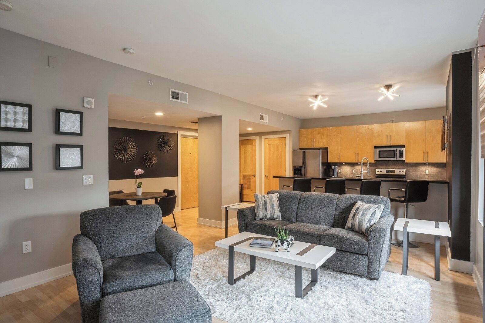 A modern living room with grey sofas, a white coffee table, and a dining area with a small table and chairs. The kitchen is visible with wooden cabinets and modern appliances.