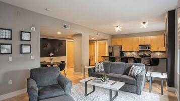 A modern living room with grey sofas, a white coffee table, and a dining area with a small table and chairs. The kitchen is visible with wooden cabinets and modern appliances.