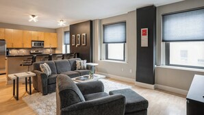 A modern apartment with a grey sofa set, a dining area with a kitchen in the background, and large windows with blinds.