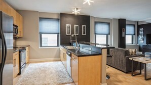 A modern kitchen with a central island, wooden cabinets, and a built-in dishwasher. There are two windows with blinds, a gray sofa, and a flat-screen TV.
