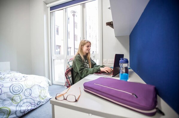 Een hotelkamer met een bed, een bureau met een laptop, een waterfles en een paarse koffer.