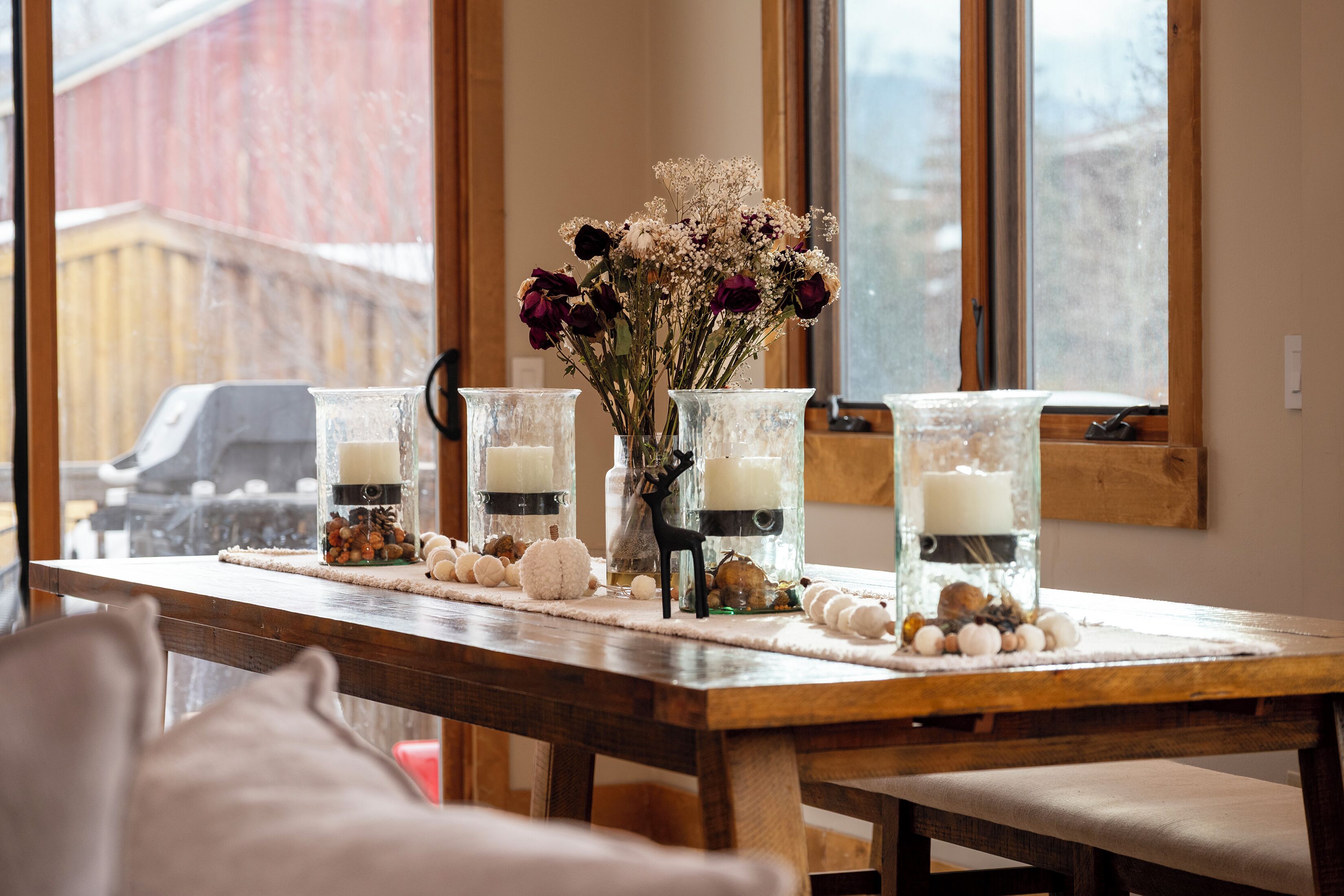 Una mesa de madera con jarrones de vidrio que contienen flores y velas, frente a una ventana con vista a un edificio rojo.