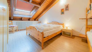 Camera con soffitto a falde, travi in legno, un letto, una sedia, un comodino con una lampada e un termosifone.