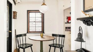 Un comedor con una mesa redonda, cuatro sillas, una ventana con vistas, una lámpara y un estante con objetos decorativos.