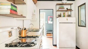 Una cocina con estufa a gas, lavamanos y refrigerador blanco. Hay libros en estanterías y un cuadro en la pared.