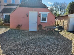 Ein kleines, pinkfarbenes Gebäude mit einer weißen Tür und einem Fenster, einem Tisch mit zwei Stühlen und einem Schotterweg.