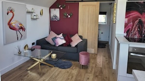 A modern living room with a dark grey sofa, a glass coffee table, and a pink ottoman. There is a kitchen visible in the background with a white oven and a marble backsplash. - Beautiful Self Contained Cottages in Westbury (Trowbridge)