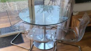 A glass-top round dining table with four clear chairs, a wooden floor, and a view of a garden through a sliding glass door. - Beautiful Self Contained Cottages in Westbury (Trowbridge)