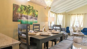 A dining area with a table set for four, a chandelier, and a painting of a waterfall on the wall. - 15min to Ocho Rios-Family sized 2bed/2bath-Pool-BBQ Grill-100Mbps-Gated Complex (Drax Hall)