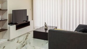 Ein modernes Wohnzimmer mit einem Flachbildfernseher auf einem schwarzen Standfuß, einem Marmorboden, einem grauen Sofa und einem großen Fenster mit transparenten Vorhängen.