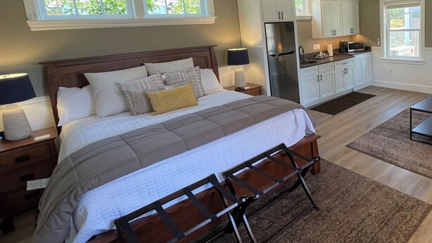 Un dormitorio con cama, mesitas de noche, lámpara, cómoda y una zona de cocina con armarios y electrodomésticos.