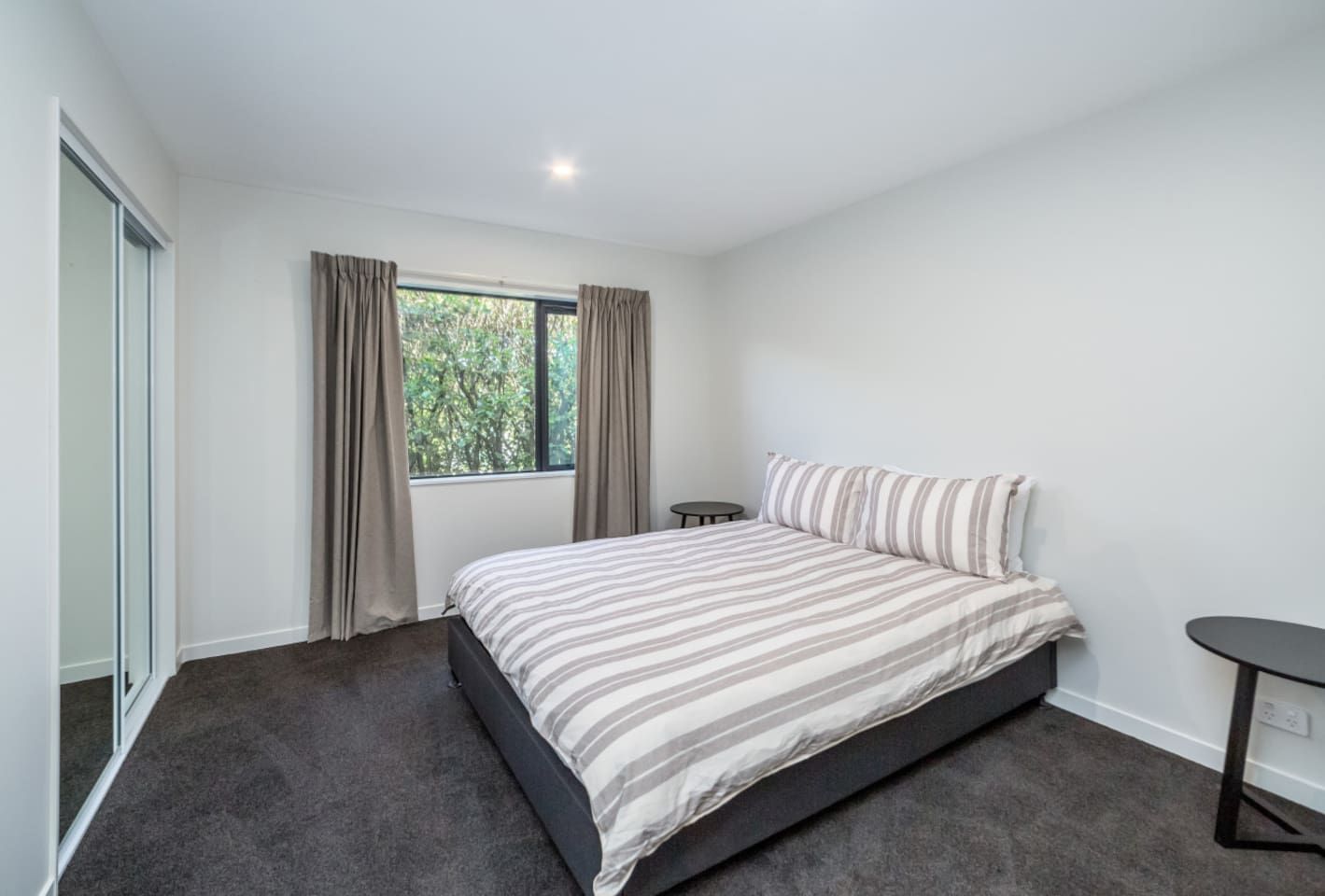 A bedroom with a bed, a small bedside table, a window with curtains, and a view of greenery outside.