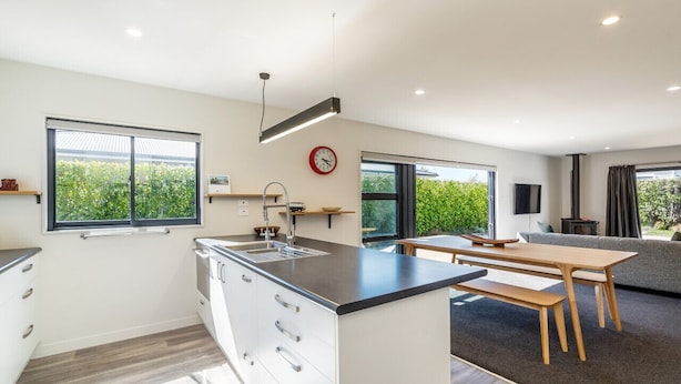 A modern kitchen with a central island, a dining area with a wooden table and benches, and large windows offering views of greenery. - Private Escape – Seasonal Communal Pool, Tennis & Relaxed Living in Pinot Grove (Martinborough)