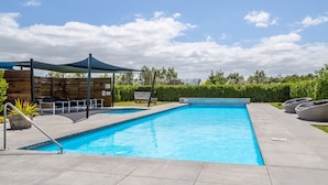 A rectangular swimming pool with a shaded seating area and lounge chairs. - Private Escape – Seasonal Communal Pool, Tennis & Relaxed Living in Pinot Grove (Martinborough)