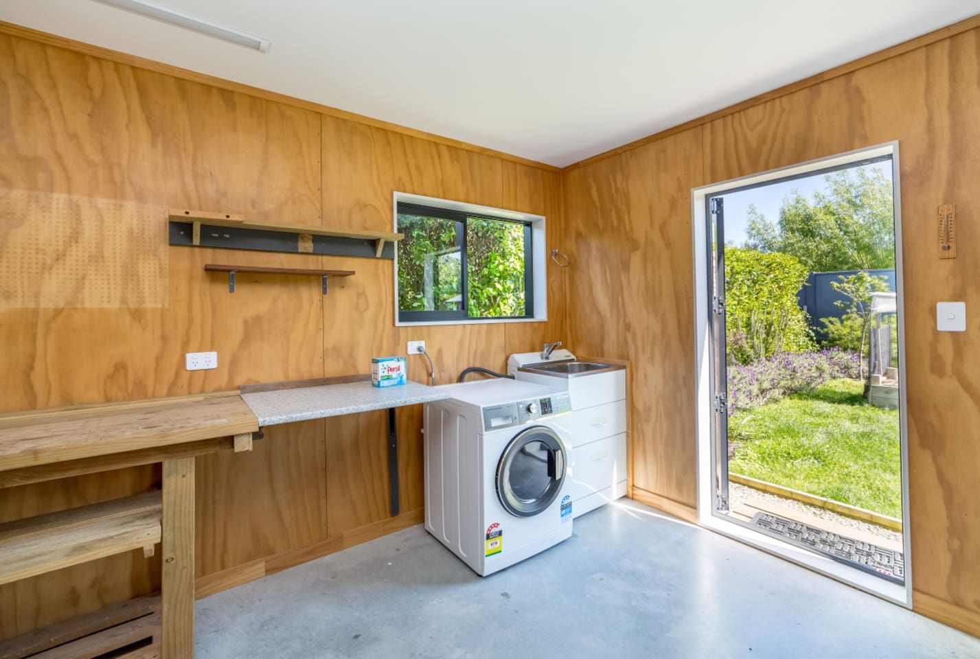 A compact laundry room with a washing machine, a sink, and a window overlooking a garden.