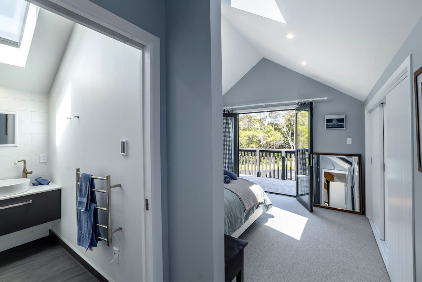A modern bedroom with a large window, a balcony, and a bathroom with a sink and towel rack.