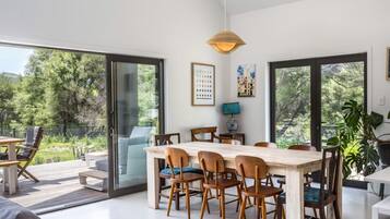 Un espace de repas moderne avec une grande table et des chaises en bois, de grandes fenêtres donnant sur un paysage verdoyant et un luminaire suspendu.
