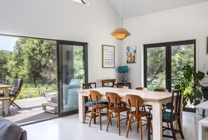 A modern dining area with a large wooden table and chairs, large windows with a view of greenery, and a hanging pendant light.