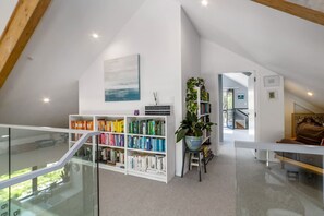 A spacious loft with a high ceiling, a large bookshelf filled with books, a potted plant, and a view of the outdoors through a glass railing. - Farmhouse Retreat, Longbush (Hinakura)