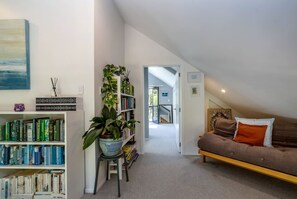 A cozy living room with a bookshelf, a large potted plant, a brown sofa with orange cushions, and a painting on the wall.