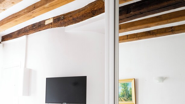 A room with a bed, a desk with chairs, a television mounted on the wall, and a painting.